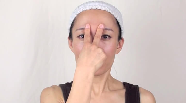 Fumiko Takatsu in a white shower cap looking straight at the camera and holding two middle fingers on her eyebrows.