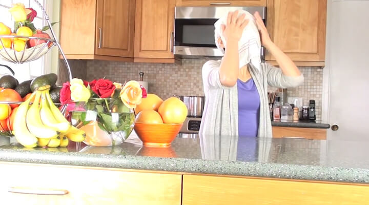 A person in the kitchen holding a white towel over her face.