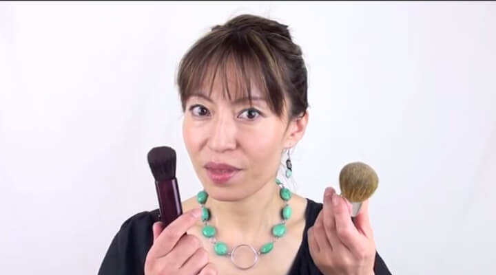 A woman with dark hair and bangs, and green necklace holding make-up brushes, one in each hand